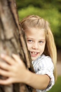 Junges Mädchen umarmt liebevoll einen Baum in die Kamera blickend