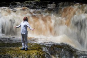 Heiliger Mann in Weiß segnet mit ausgebreiteten Armen das Wasser vor einem Wasserfall stehend