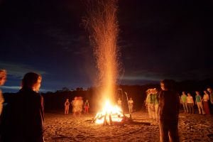 Bild eines großen loderndes Feuers mit dunkelblauem Wolkenhimmel und drumherum stehenden Menschen