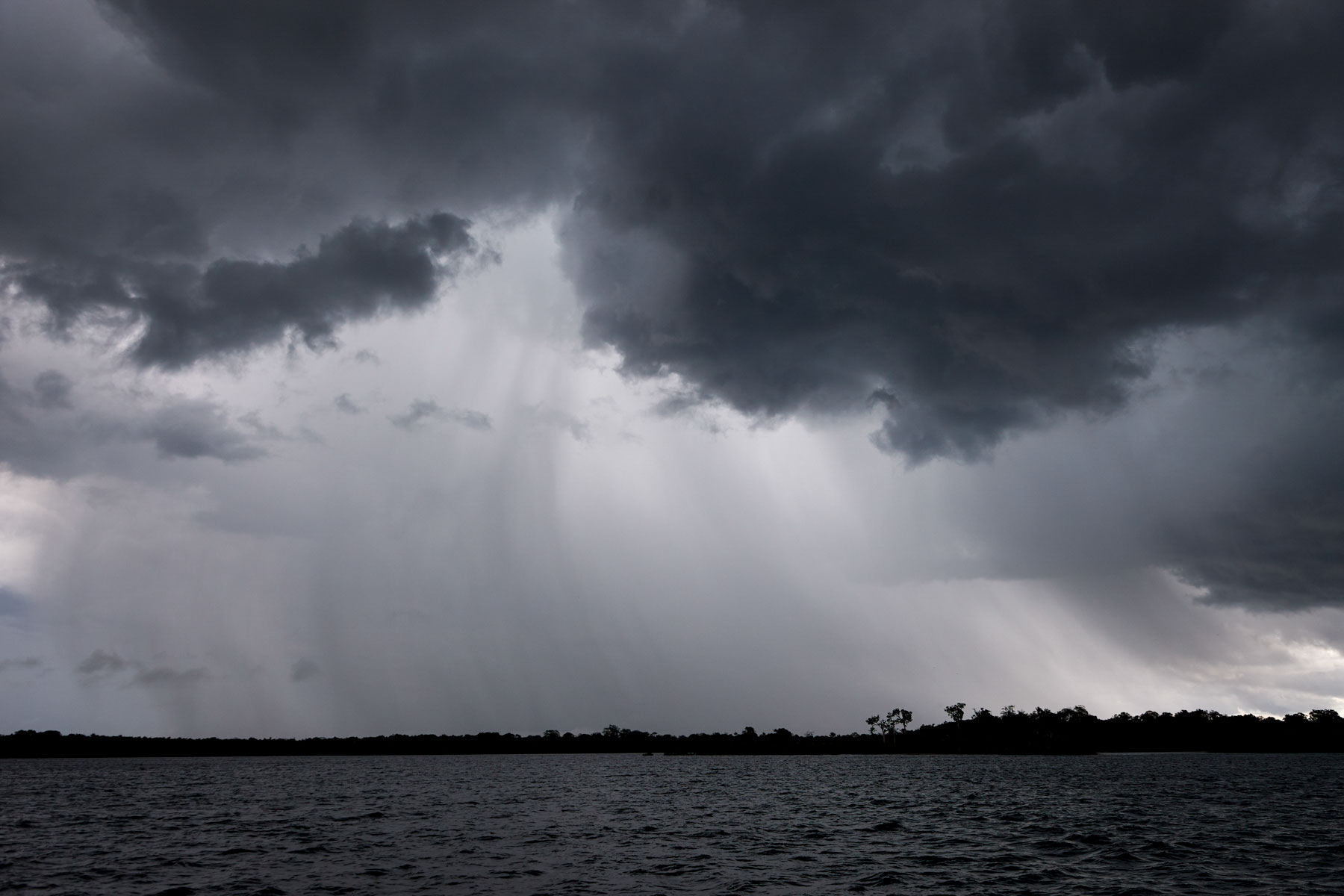 Bedrohliches Bild einer riesigen dunklen Regenwolke über einem Gewässer, aus der sich der sichtbare Regen ergießt