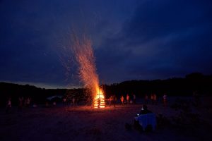 Bild eines großen Feuers mit dunkelblauem Wolkenhimmel und drumherum stehenden Menschen
