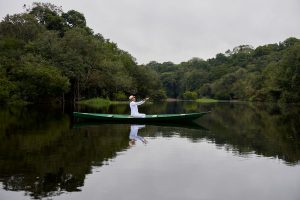Foto eines Mannes in Weiß mit ausgestreckten Armen in einem Holzboot sitzend, das Wasser und den umliegenden Regenwald segnet