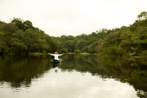 Foto eines Mannes in Weiß mit ausgebreiteten Armen in einem Holzboot sitzend, das Wasser und den umliegenden Regenwald segnet