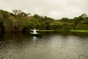 Foto eines Mannes in Weiß in einem Holzboot sitzend, das Wasser und den umliegenden Regenwald segnet