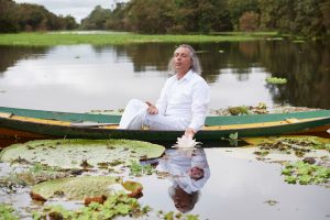 Heiliger Mann in weiß, segnet in einem Holzboot sitzend mit geschlossenen Augen eine blühende, wunderschöne Seerose