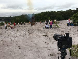 Foto einer Gruppe, die im Kreis um ein Feuer stehen mit einer Kamera im Vordergrund