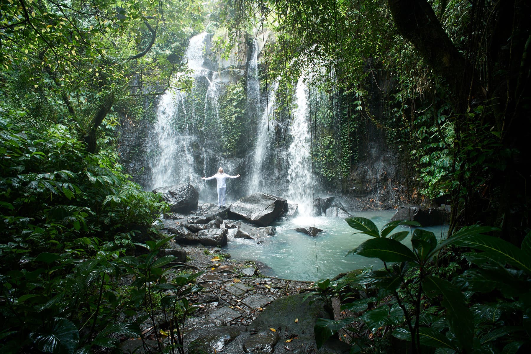 Bild zeigt einen paradiesischen Wasserfall im Dschungel in dem eine Person segnend die Arme ausbreitet