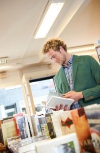 Portrait eines Mannes in hellgrünem Jacket in einer Buchhandlung, der in einem Buch blättert