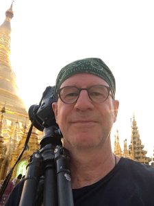 Mann mit geschulterter Kamera auf Stativ schaut vor einem Tempel in die Kamera