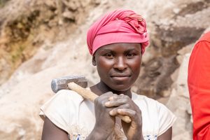 Portrait einer jungen afrikanischen Frau mit rotem Kopftuch und geschultertem Schlaghammer im Steinbruch